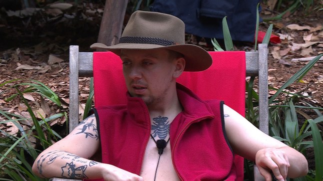Aitch sits in a red chair wearing a hat and a zip-up fleece.