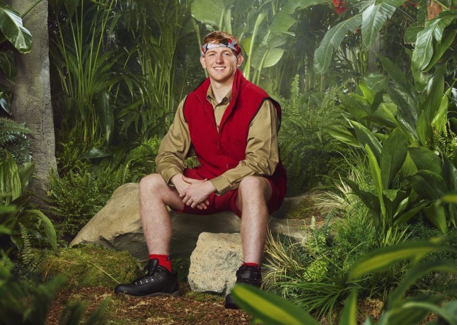 Angry Ginge smiling in a jungle photoshoot for I'm A Celebrity, wearing a red vest and beige shirt.